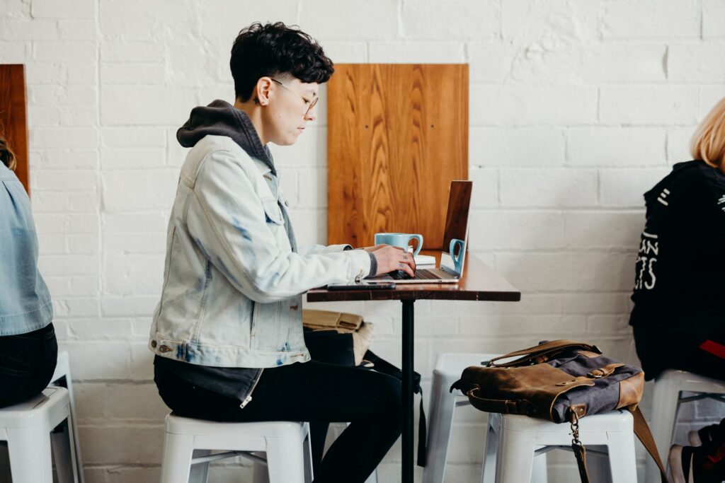 Better work communication is possible anywhere, seen in this person using a laptop in a cafe with their messenger bag on the stool in front of them.