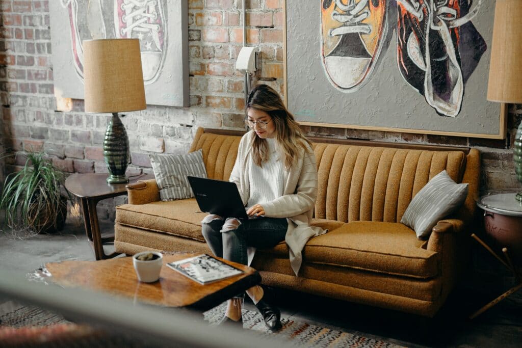 A person sat on a sofa using a laptop in a room with large paintings behind them on the wall as they learn how to run a hybrid office.