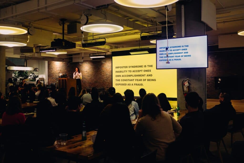 A person stood in front of an audience giving a presentation in a large room standing next to a projected screen knowing how to make a pitch deck.