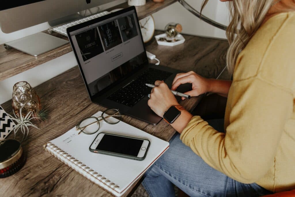 A person sat at their desk at home working on a laptop with a notebook, glasses and a phone on a desk looking for the best work from home apps.