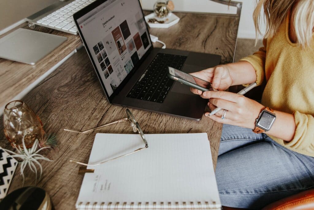 Someone holding their phone, sitting down, while using a laptop on their desk with a notepad on the table and glasses on top of that while working from home.