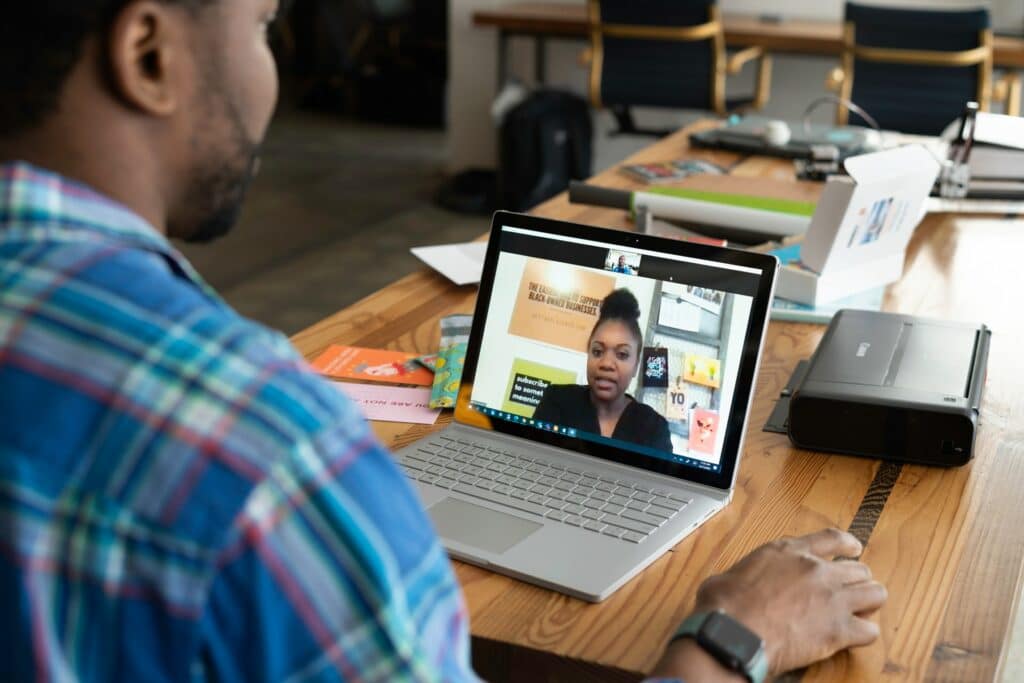 A person attending a virtual meeting with one other person on their laptop screen using a long desk with a printer and many other items on it.