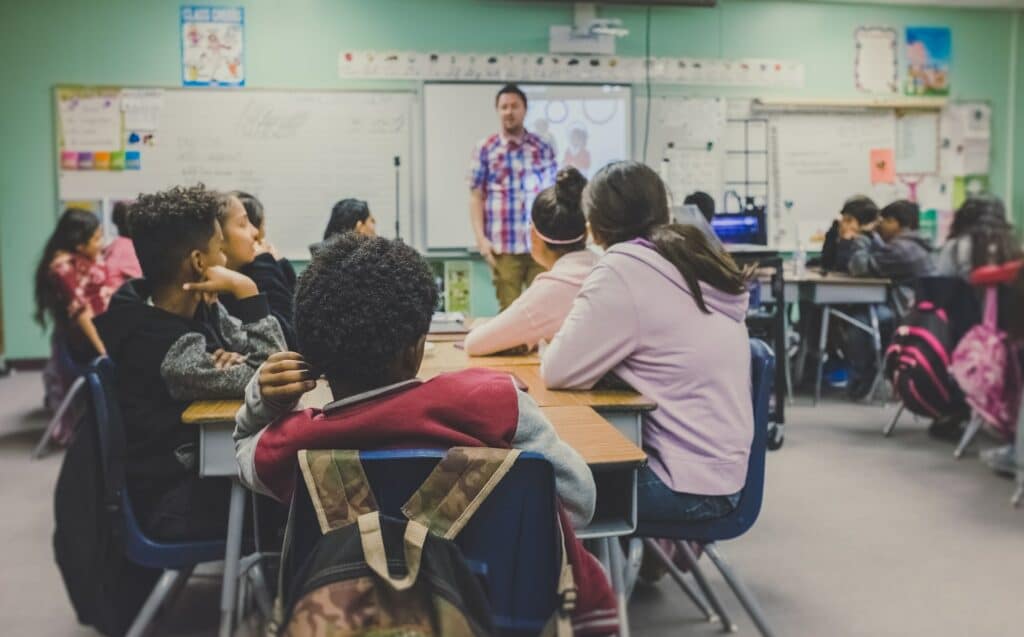 Children in a classroom all looking at a teacher who is using an old projector to show the class what they're learning, using XSplit Presenter in this scenario is a good way to improve teaching.