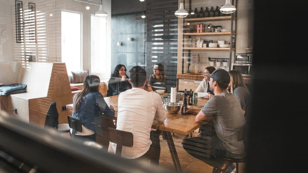 A meeting room setting with seven people sat arouynd a table discussing their work as part of a hybrid office.